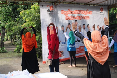 A group of people wearing colorful headscarves and shirts are standing outdoors with their arms raised, seemingly participating in an exercise or stretching activity. They are in front of a large banner featuring images and text. The setting includes greenery and a covered area.