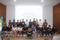 A group of people is posing for a photograph in a room with a white screen in the background that has text welcoming e-sport participants for a Mobile Legends event organized by PKS Prambanan. The group consists of 20 individuals standing and sitting, with some wearing casual attire and others wearing matching shirts that include logos.
