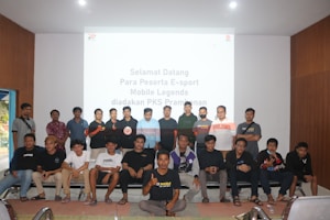 A group of people is posing for a photograph in a room with a white screen in the background that has text welcoming e-sport participants for a Mobile Legends event organized by PKS Prambanan. The group consists of 20 individuals standing and sitting, with some wearing casual attire and others wearing matching shirts that include logos.