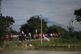 A group of people wearing orange and white shirts are cycling and walking along a rural road surrounded by greenery. A truck with people and flags is also visible. In the background, a mountain rises under a cloudy sky, and there are utility poles and trees lining the road.