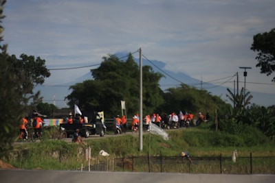 A group of people wearing orange and white shirts are cycling and walking along a rural road surrounded by greenery. A truck with people and flags is also visible. In the background, a mountain rises under a cloudy sky, and there are utility poles and trees lining the road.