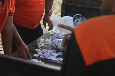 A group of people in orange shirts gather around a table covered with white circular pins. Some pins are enclosed in plastic packaging, while others are scattered around. One person is pointing at the pins, and another is reaching out to pick them up.