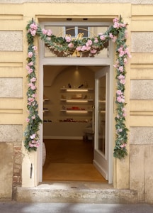 A charming storefront entrance adorned with pink and green floral garlands framing the door. The interior visible through the open glass door shows shelves with various merchandise including handbags and shoes, creating an inviting boutique atmosphere.