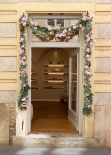 A charming storefront entrance adorned with pink and green floral garlands framing the door. The interior visible through the open glass door shows shelves with various merchandise including handbags and shoes, creating an inviting boutique atmosphere.