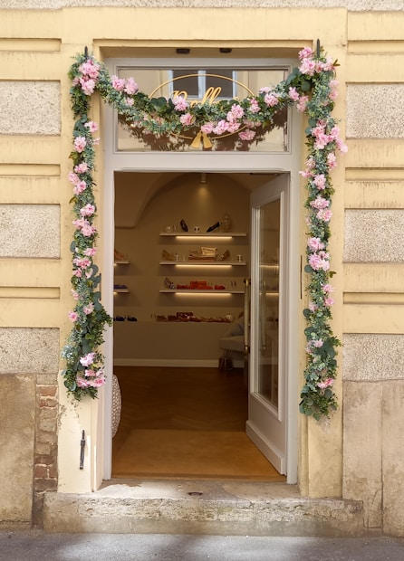 A charming storefront entrance adorned with pink and green floral garlands framing the door. The interior visible through the open glass door shows shelves with various merchandise including handbags and shoes, creating an inviting boutique atmosphere.