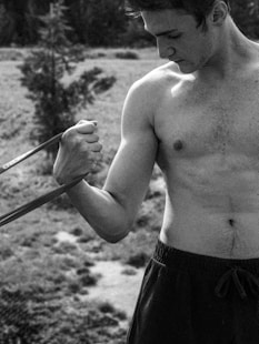 A shirtless man outdoors exercises with a resistance band, focusing on his bicep. The background is a blurred natural setting with grass and trees, suggesting a park or forest environment.