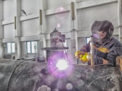 A person wearing protective gear is welding a large metal pipe indoors. Sparks and bright light emanate from the welding process, with the individual holding the welding apparatus with one hand while steadying it with the other. The workspace has industrial features, with walls, windows, and various machinery visible.