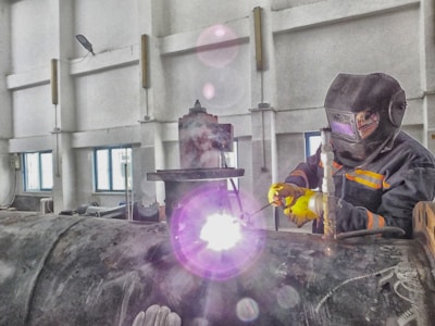 A person wearing protective gear is welding a large metal pipe indoors. Sparks and bright light emanate from the welding process, with the individual holding the welding apparatus with one hand while steadying it with the other. The workspace has industrial features, with walls, windows, and various machinery visible.