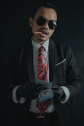 A man wearing a black suit, white shirt, and a red paisley tie is holding a stack of money. He is also wearing black gloves and dark sunglasses. A cigar is clenched between his teeth.