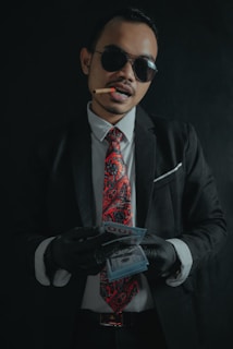 A man wearing a black suit, white shirt, and a red paisley tie is holding a stack of money. He is also wearing black gloves and dark sunglasses. A cigar is clenched between his teeth.