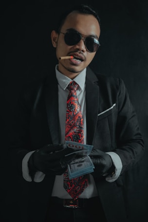 A man wearing a black suit, white shirt, and a red paisley tie is holding a stack of money. He is also wearing black gloves and dark sunglasses. A cigar is clenched between his teeth.