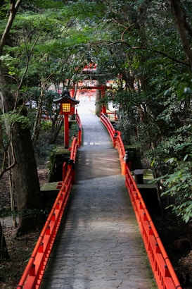A tranquil stone path flanked by vibrant red railings winds through lush greenery. Traditional Japanese lanterns are placed along the path, providing a serene and culturally rich atmosphere. The path leads to a traditional torii gate, partially obscured by the foliage that surrounds it, suggesting a feeling of exploration and serenity.