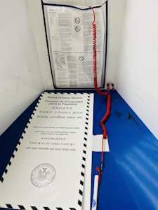 A blue voting booth contains instructions in multiple languages. A privacy sleeve with a government seal is on a ledge, and a pen with a red curly cord is attached nearby.
