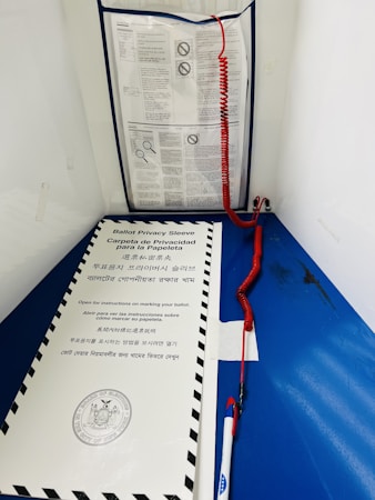 A blue voting booth contains instructions in multiple languages. A privacy sleeve with a government seal is on a ledge, and a pen with a red curly cord is attached nearby.