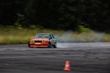 A car is drifting on a racetrack, producing smoke as it moves. The vehicle is an older model with a red front and black detailing. An orange traffic cone is visible on the track, and the background features a blurred, forested area, indicating motion.
