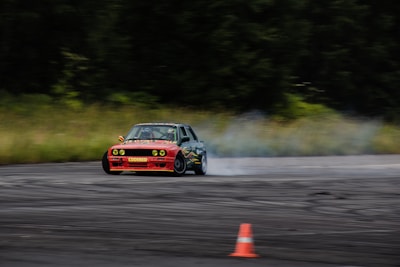 A car is drifting on a racetrack, producing smoke as it moves. The vehicle is an older model with a red front and black detailing. An orange traffic cone is visible on the track, and the background features a blurred, forested area, indicating motion.