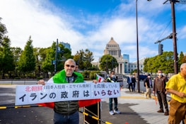 A group of people are gathered outdoors near a government building, holding signs with Japanese and other languages. One individual in the foreground wears sunglasses and a jacket, visibly holding a sign. The scene is set on a bright day with a clear blue sky. Trees and well-maintained gardens surround the area.