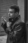 A man wearing a leather jacket stands next to a building, holding his collar with one hand while appearing to smoke. The backdrop includes a brick wall and horizontal metal grating, suggesting an urban environment. The image is captured in black and white, highlighting textures and shadows.