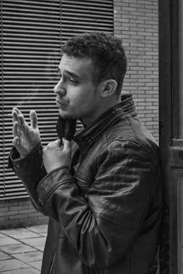 A man wearing a leather jacket stands next to a building, holding his collar with one hand while appearing to smoke. The backdrop includes a brick wall and horizontal metal grating, suggesting an urban environment. The image is captured in black and white, highlighting textures and shadows.