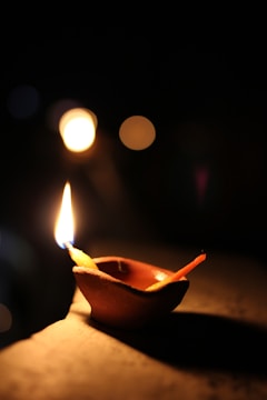 A small clay lamp with a lit flame stands on a surface, illuminating its surroundings with a soft glow. The background is dark, creating a stark contrast with the warm light of the lamp. Several out-of-focus lights appear as blurred circles, contributing to a serene and contemplative atmosphere.