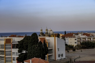 A coastal townscape with residential buildings featuring white and terracotta roofs. In the background, a body of water is visible, and two church towers with distinctive architecture rise. Cypress trees are visible in the foreground.