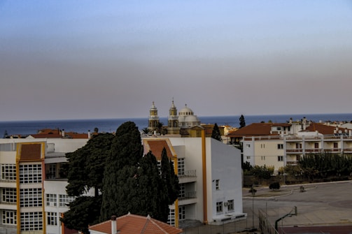 A coastal townscape with residential buildings featuring white and terracotta roofs. In the background, a body of water is visible, and two church towers with distinctive architecture rise. Cypress trees are visible in the foreground.