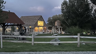 A rustic barn with illuminated windows stands amidst a lush green lawn bordered by wooden fences. The barn's structure is complemented by a wooden water tower nearby. Large trees surround the area, and a small open shelter with a roof is visible on the left. The sky is overcast, suggesting an early evening or near-storm atmosphere.