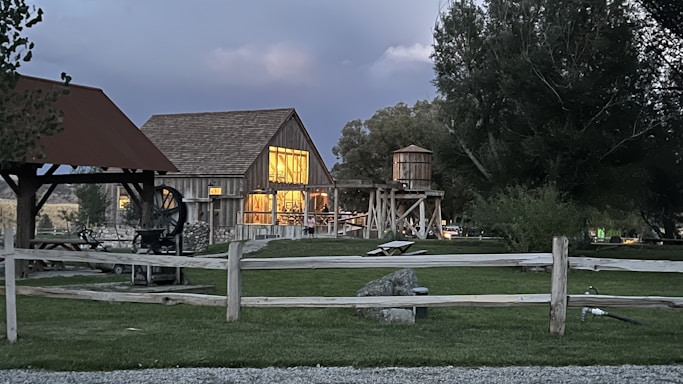 A rustic barn with illuminated windows stands amidst a lush green lawn bordered by wooden fences. The barn's structure is complemented by a wooden water tower nearby. Large trees surround the area, and a small open shelter with a roof is visible on the left. The sky is overcast, suggesting an early evening or near-storm atmosphere.