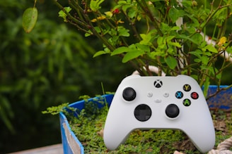A white gaming controller with black buttons and colorful highlights rests on a patch of green plants in a blue container. The background features lush green foliage, contributing to a natural setting.