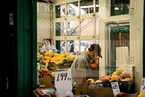 A small produce shop features a variety of fresh fruits and vegetables neatly arranged. Yellow squash and orange pumpkins are prominently displayed alongside bags of potatoes. A person wearing a cap and mask is tending to the produce inside the shop. Several price tags are visible, indicating the prices of strawberries and other fruits. The shop has glass windows, allowing natural light to illuminate the interior.