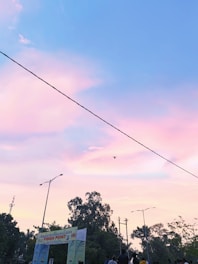 The image features a clear sky transitioning from blue to pink and purple hues during twilight. A long, thin wire stretches diagonally across the frame, with a drone flying nearby. In the foreground, the silhouettes of trees and lampposts are visible, along with a colorful banner hanging between two posts, indicating a public event or gathering.