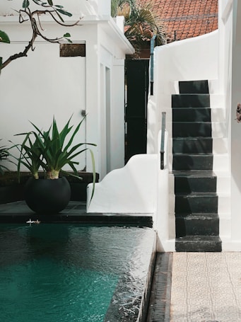 A serene outdoor area features a turquoise pool with calm water. A modern, minimalist staircase with black steps leads upwards alongside a white wall. In the background, lush green plants are placed in a large black pot next to the pool. The overall architecture is clean and contemporary, with white walls and a red-tiled roof visible through foliage.