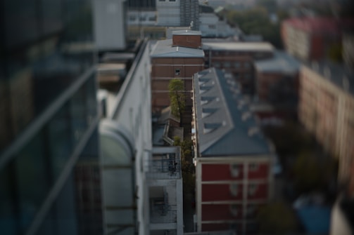 An aerial view of a city landscape features several buildings with a tilt-shift effect, causing parts of the image to appear out of focus. The architecture includes modern structures as well as older brick buildings. A narrow alleyway is visible between the buildings, and some greenery is present along the path.