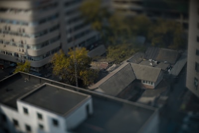Aerial view of an urban area featuring a mix of large modern buildings and smaller traditional houses. The scene is slightly blurred, creating a tilt-shift effect. Trees with green and yellow foliage add a hint of nature amidst the dense structures.