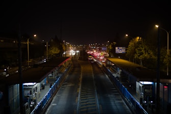 A nighttime urban setting with a busy road and traffic. Streetlights illuminate the scene, casting light on the trees and road signage. Two bus stations with shelters flank the road, and several vehicles are queued with red taillights visible.