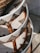 A close-up view of intricately woven rattan furniture with a geometric pattern. The craftsmanship features a combination of natural brown and white fibers, enhanced by sunlight. The image captures the texture and design of the layered wicker material.