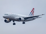 A commercial airplane bearing the Croatia Airlines branding is captured in mid-flight against a cloudy gray sky. The aircraft has a sleek white body with distinctive red and blue checkered patterns on the vertical stabilizer.