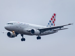 A commercial airplane bearing the Croatia Airlines branding is captured in mid-flight against a cloudy gray sky. The aircraft has a sleek white body with distinctive red and blue checkered patterns on the vertical stabilizer.