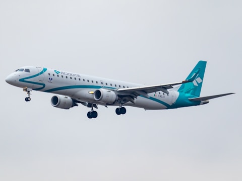 A commercial airplane with a predominantly white body and teal accents flies in the sky. The aircraft is a twin-engine jet with its landing gear extended, indicating it is likely approaching for landing. The overcast sky gives a muted backdrop.