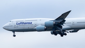 An aircraft featuring the Lufthansa airline logo is in flight, with its landing gear extended. The plane's body is mostly white, and it is flying against a backdrop of an overcast sky.