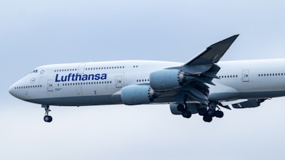 An aircraft featuring the Lufthansa airline logo is in flight, with its landing gear extended. The plane's body is mostly white, and it is flying against a backdrop of an overcast sky.