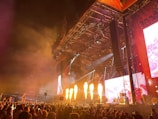 A large crowd gathers in front of a concert stage during a night performance. Bright flames shoot up from the stage, enhancing the lively atmosphere. The stage is equipped with large screens displaying visuals, and vibrant lighting creates dynamic patterns. Tall structures and speakers frame the stage, while the audience raises their hands and flags in excitement.