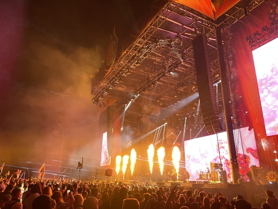 A large crowd gathers in front of a concert stage during a night performance. Bright flames shoot up from the stage, enhancing the lively atmosphere. The stage is equipped with large screens displaying visuals, and vibrant lighting creates dynamic patterns. Tall structures and speakers frame the stage, while the audience raises their hands and flags in excitement.