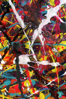 An abstract painting featuring a chaotic mixture of vibrant colors such as red, yellow, blue, and black. The composition includes random and energetic splatters, drips, and streaks of paint, creating a dynamic and intense visual experience. Thick white streaks intersect and contrast with the multicolored background.