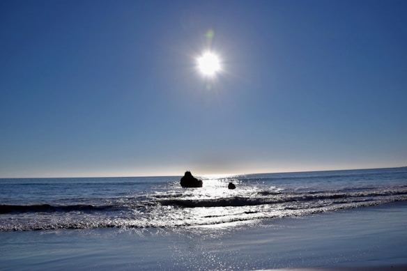The image captures a serene beach scene with a bright sun shining in the sky, casting sparkling reflections on the ocean water. The horizon is clear and visible, and two rocks are seen protruding from the sea, adding to the tranquil coastal atmosphere.