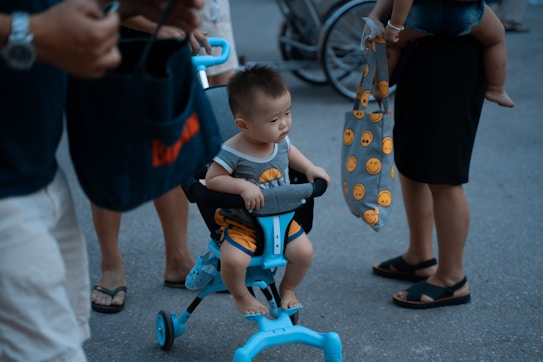 A toddler sits in a turquoise and black tricycle with a seat, surrounded by several adults standing nearby. One adult holds a gray bag with bright yellow emoji faces. The scene takes place on a street or outdoor area with a bicycle visible in the background.