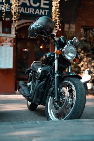 A black motorcycle is parked in front of a restaurant with a sign partially visible above. The motorcycle is sleek and shiny, equipped with a helmet resting on the handlebar. The scene is urban with a warm ambiance, enhanced by hanging plants and the reflection of outdoor lights.