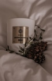 A white candle with a gold label reading 'Almond Pumpkin Limited Edition', surrounded by leaves and pinecones, resting on a soft, light-colored fabric, creating a cozy and warm atmosphere.