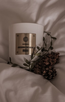 A white candle with a gold label reading 'Almond Pumpkin Limited Edition', surrounded by leaves and pinecones, resting on a soft, light-colored fabric, creating a cozy and warm atmosphere.