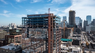 A cityscape featuring a high-rise building under construction, with cranes visible on a clear day. The partially built structure is surrounded by other completed skyscrapers and urban architecture, indicating a rapidly developing city area.